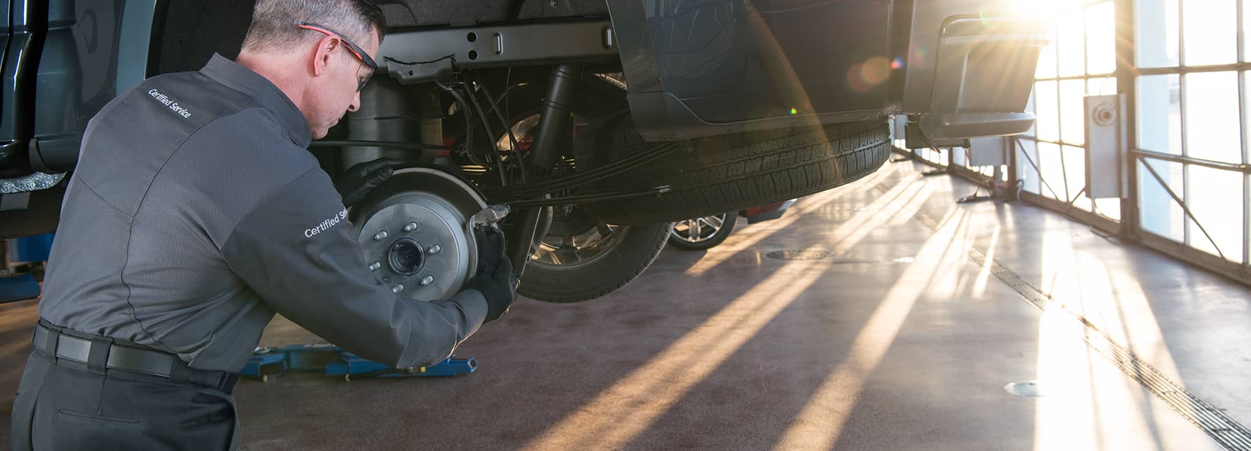 Mechanic performing brake service on a lifted vehicle in a sunlit garage.