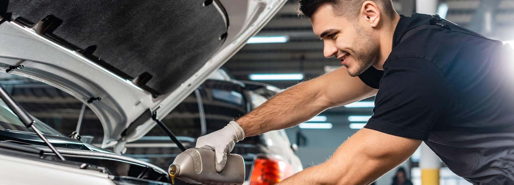 Mechanic smiling while pouring oil into a car engine in a garage setting.