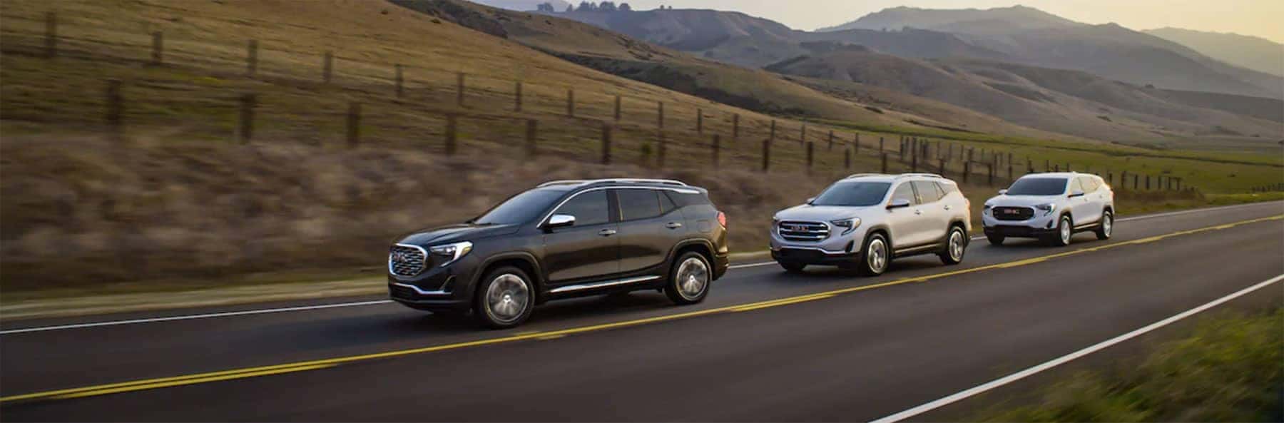 Three SUVs driving on a rural highway at sunset, with hills in the background.