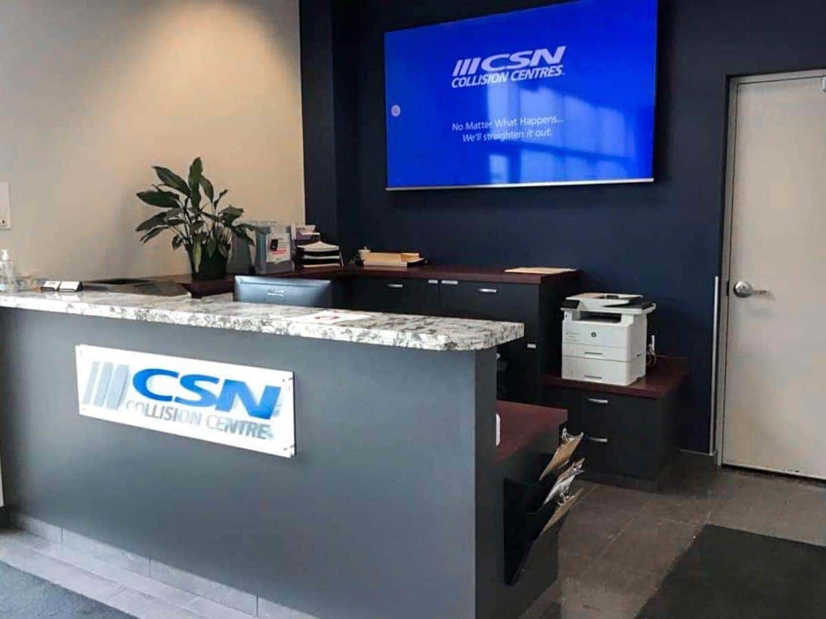 Reception area of CSN Collision Centre with countertop, plant, printer, and wall-mounted screen.
