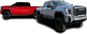Two pick-up trucks, one red and one gray, are parked on a runway or a large parking lot. The red truck is at an angle that makes it look farther away and smaller, while the gray truck is closer and larger. Behind them, a city skyline with tall buildings can be seen under a blue sky with scattered cloud