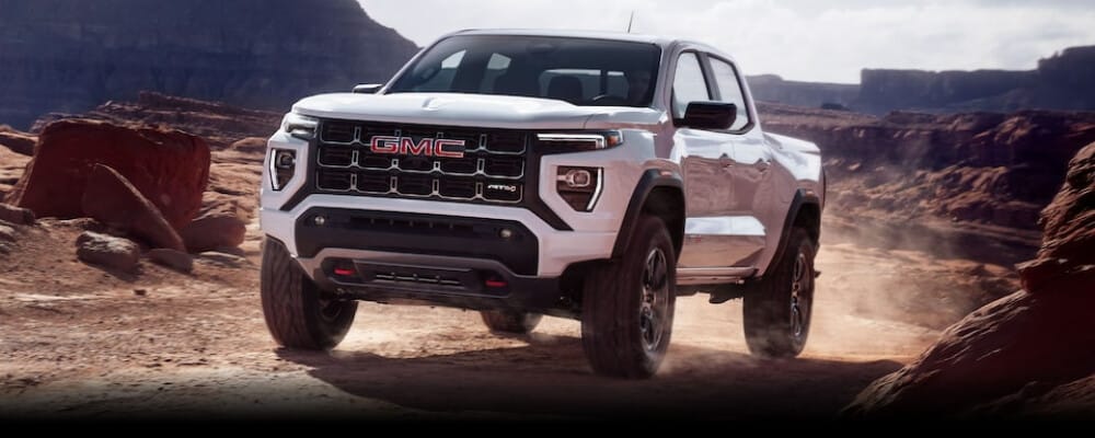 White GMC AT4 truck driving on a rugged desert landscape with red rocks and distant cliffs.