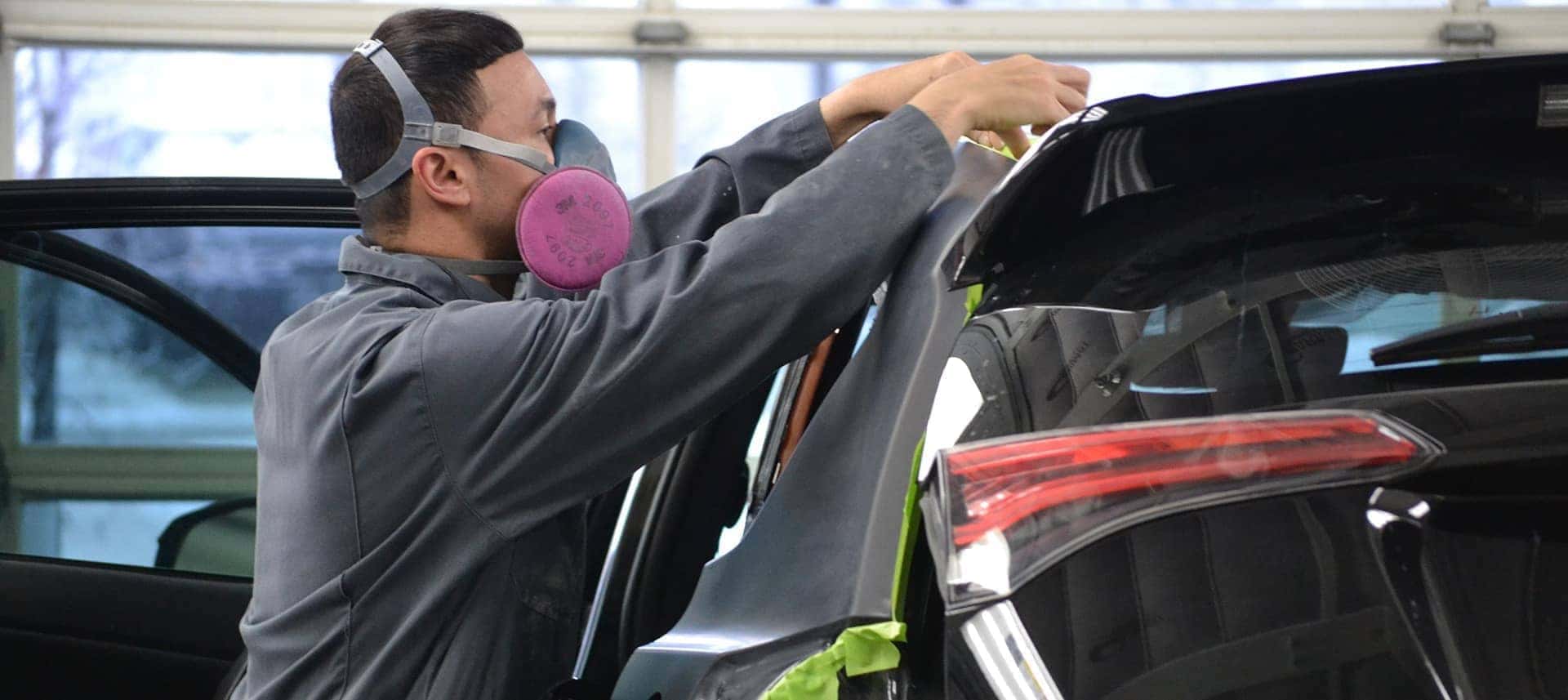 Auto technician wearing a respirator working on a car's rear window repair.