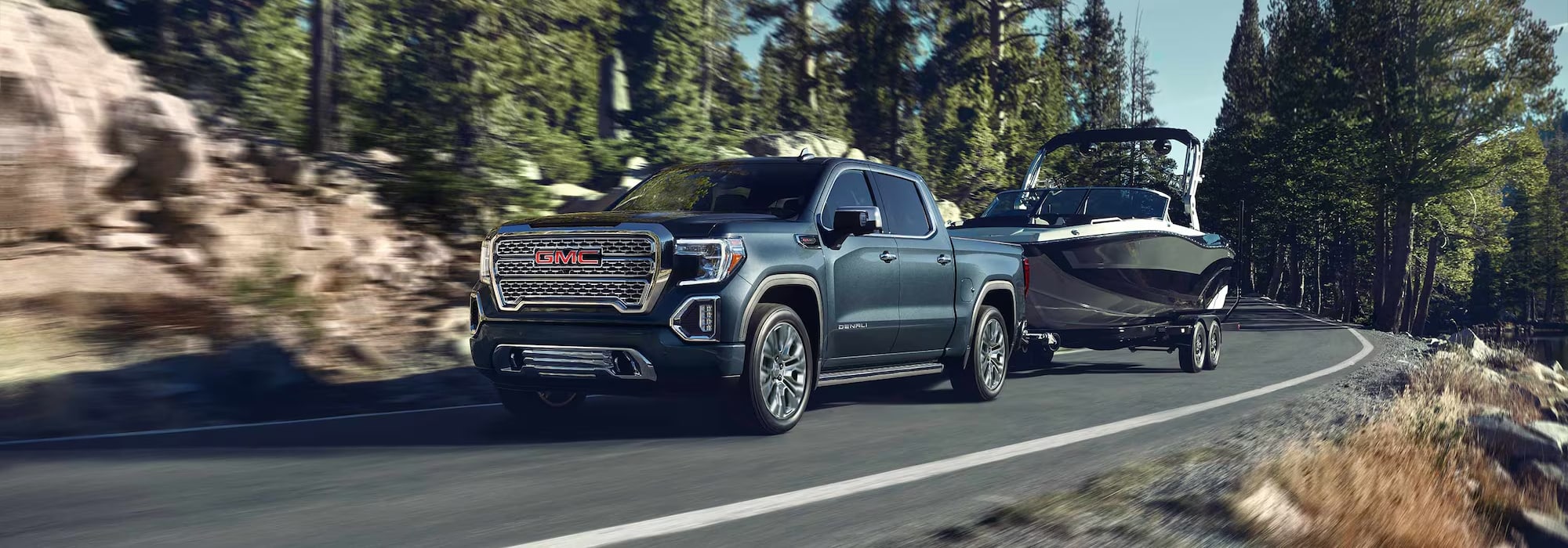 A GMC truck towing a boat on a scenic mountain road with lush forests and rocky terrain.