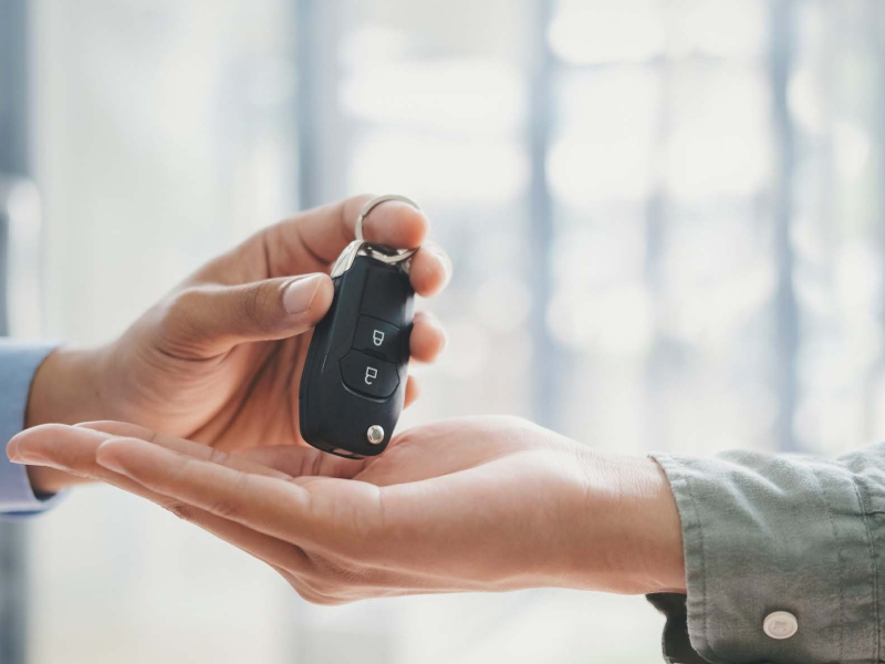 Hand giving car key to another hand, symbolizing car ownership or rental transaction.