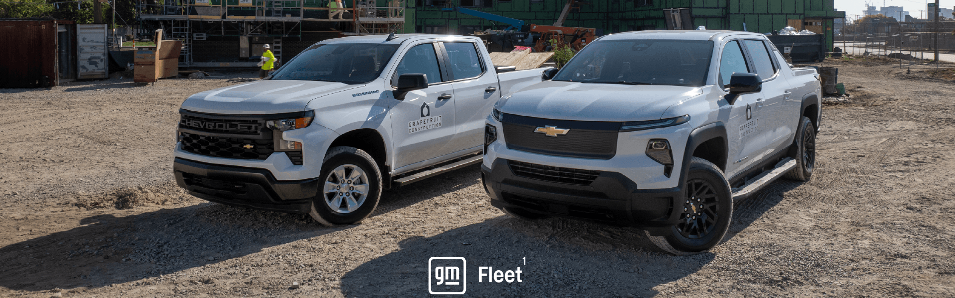 Two Chevrolet pickup trucks on a construction site with buildings and workers in the background.